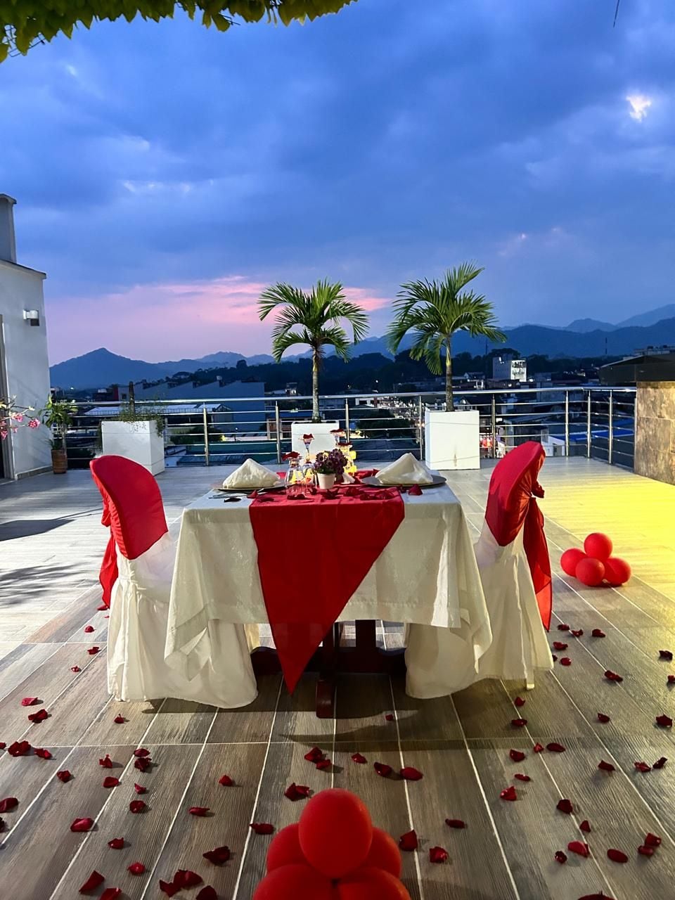 Un espace de restauration en plein air avec une table dressée pour deux personnes, entouré de chaises dotées de coussins rouges, une vue sur la mer et les montagnes, et un coucher de soleil en arrière-plan.