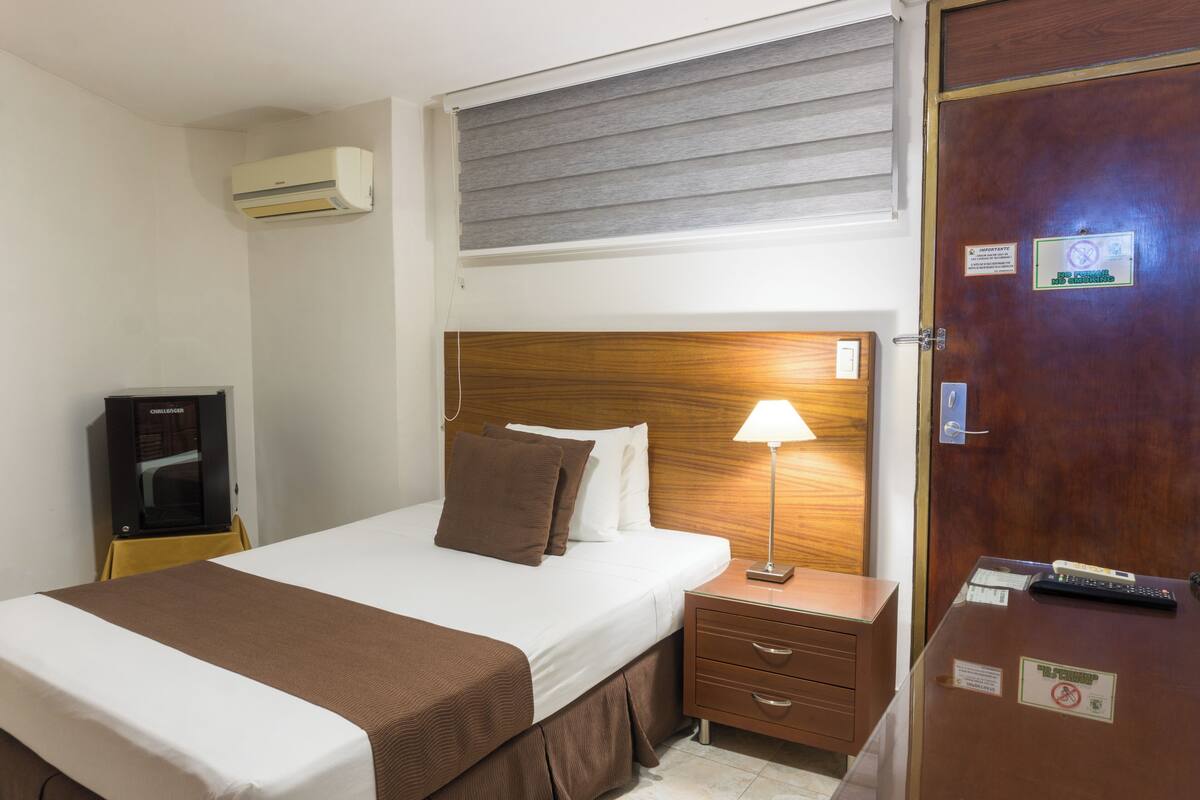 A hotel room with a bed, bedside lamp, wooden headboard, a nightstand, a desk with a remote control, and an air conditioning unit.