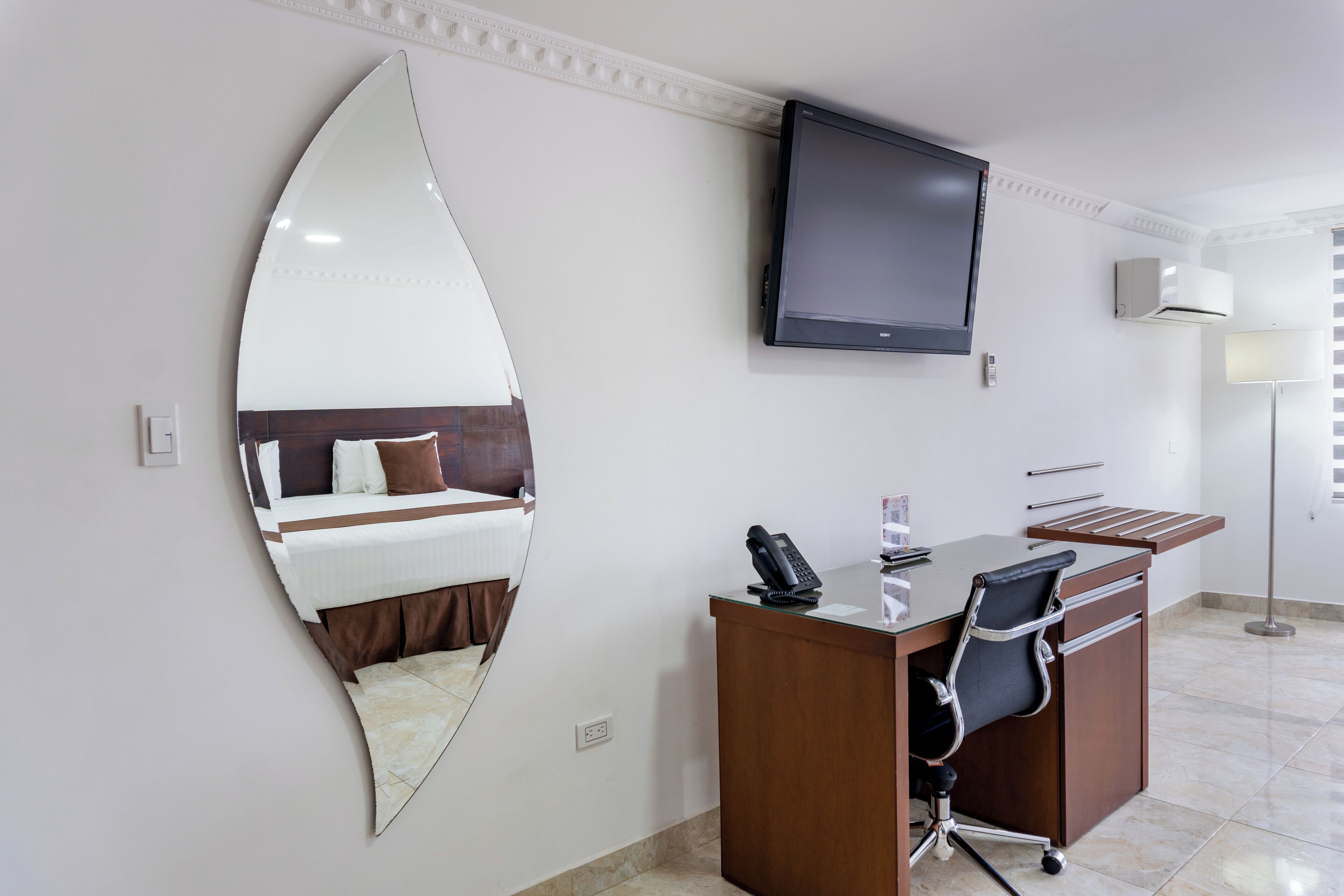 A hotel room with a bed, a desk with a telephone, a flat-screen TV, and a large mirror.