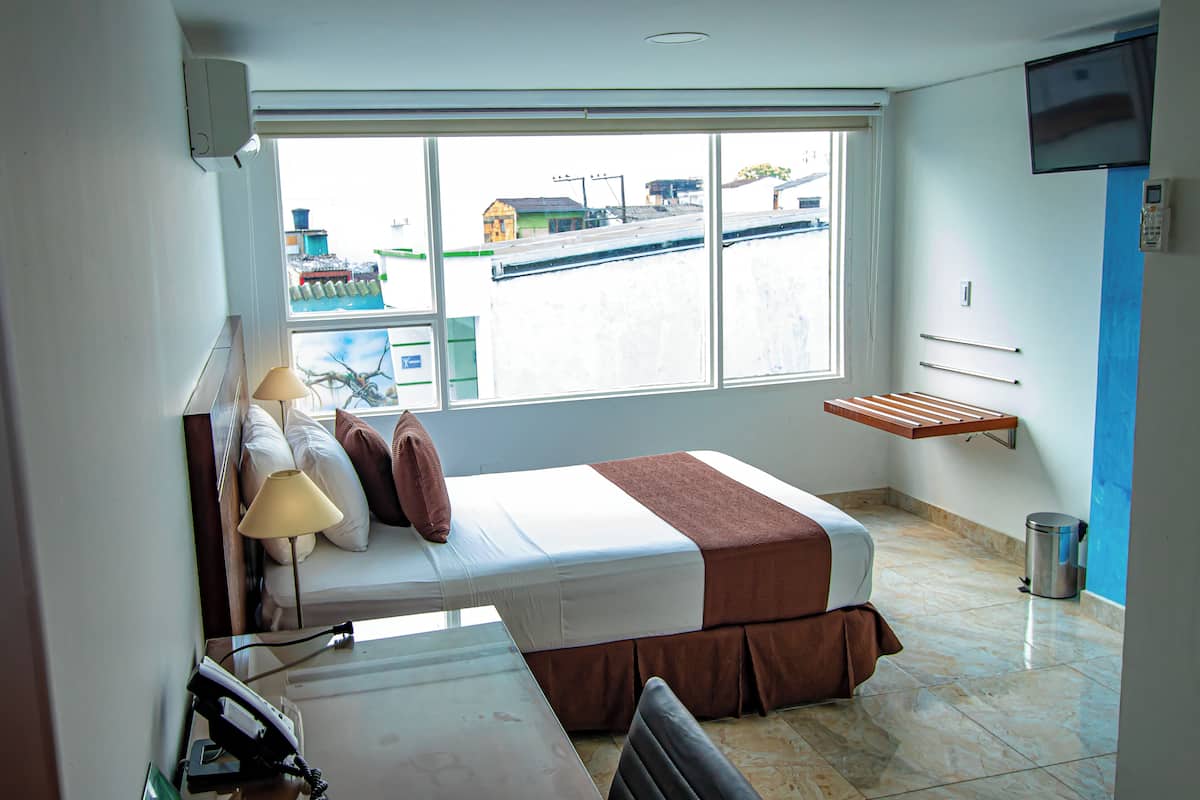 A hotel room with a bed, a desk, a chair, a lamp, a TV, a window, and a trash bin.