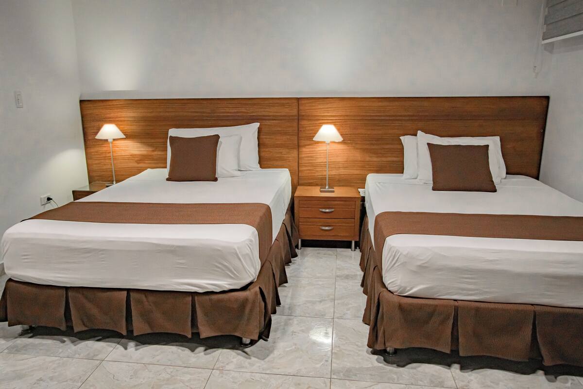 A hotel room with two beds, each with a wooden headboard, white linens, and brown accents. There are bedside tables with lamps and a small drawer unit between the beds.