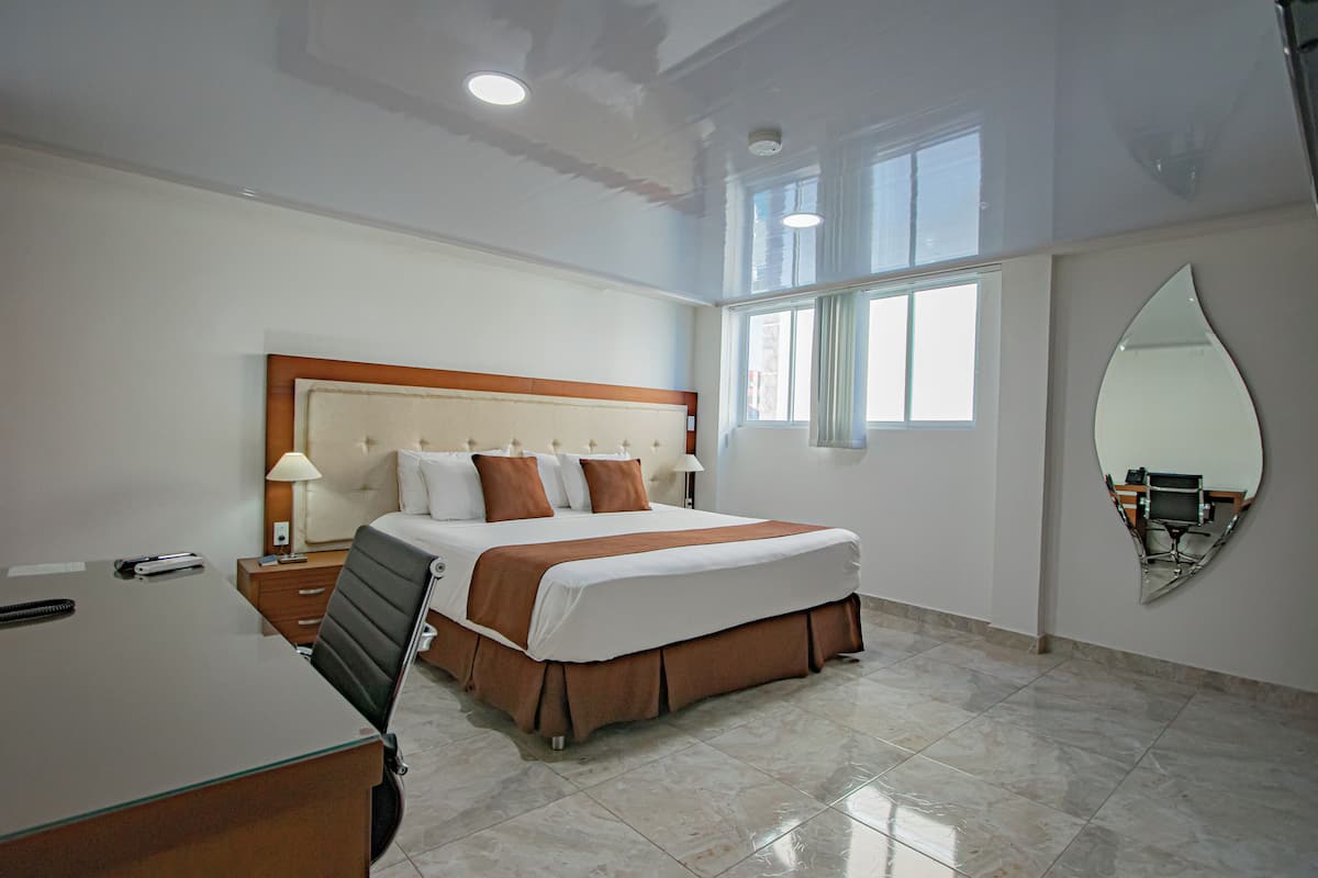 A hotel room with a large bed, a desk, a chair, a nightstand, a mirror, and a window.