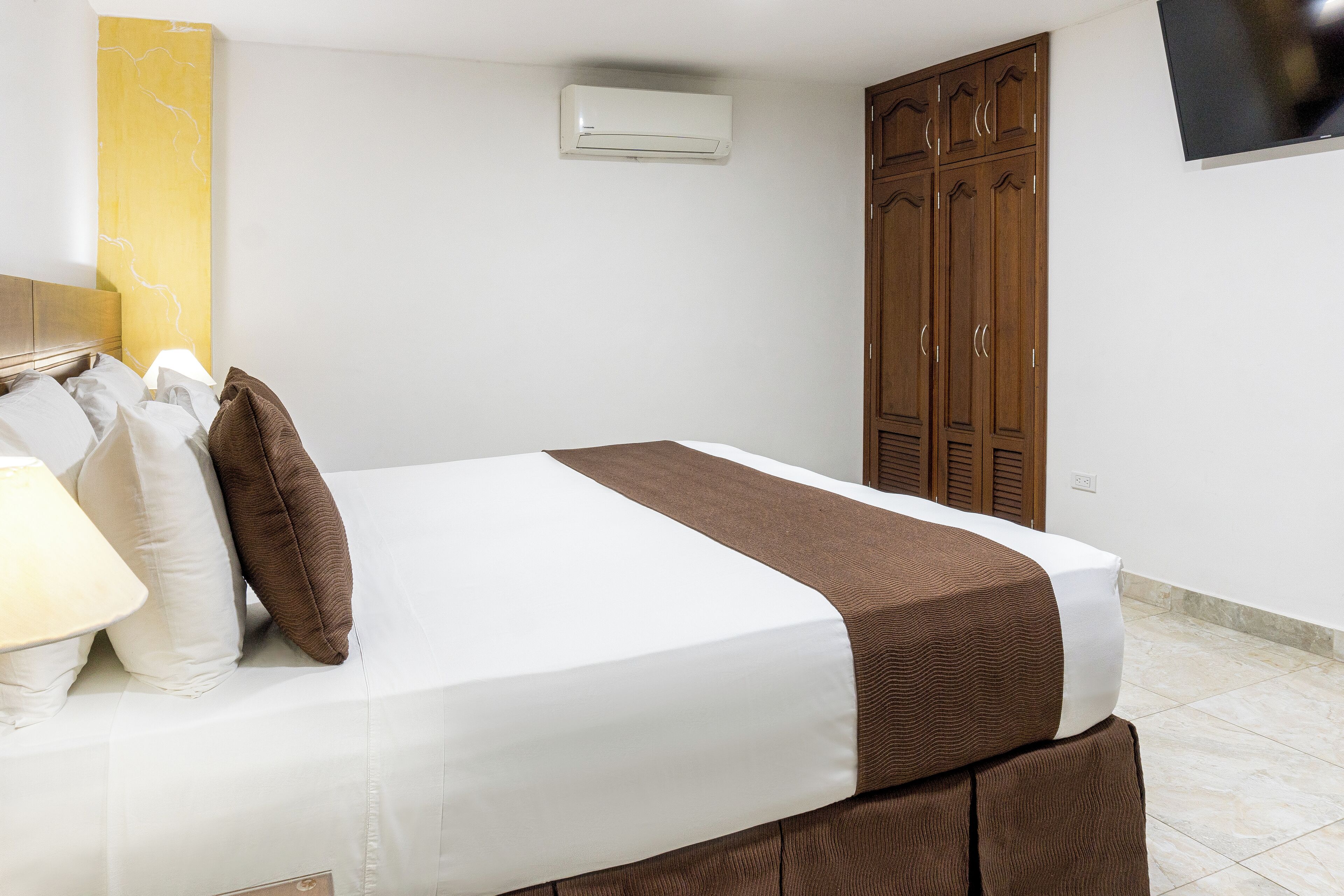 A neatly made bed with a white and brown bedspread, a bedside lamp, and a wooden wardrobe with a TV mounted on the wall.