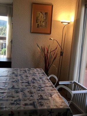 Un comedor con una mesa cubierta con un mantel de temática náutica, sillas blancas, una lámpara de pie y un cuadro en la pared.