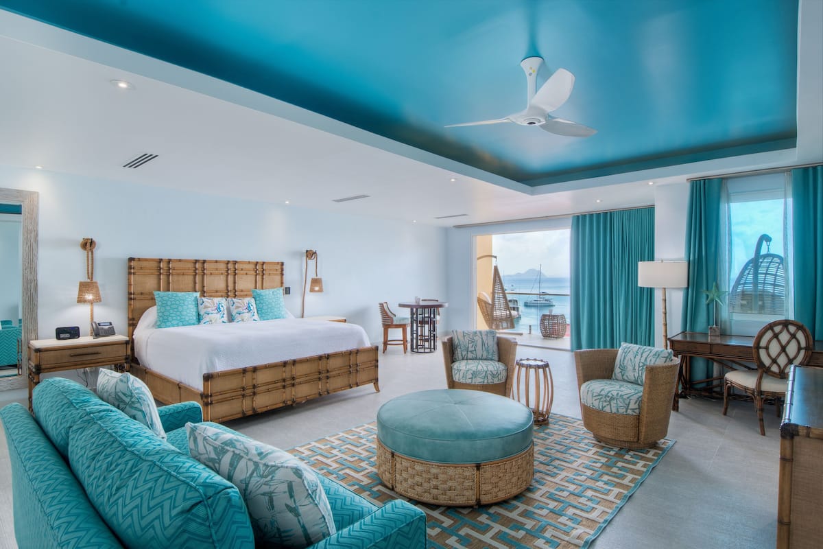 A modern hotel room with a large bed, a sofa, a round ottoman, and a desk.