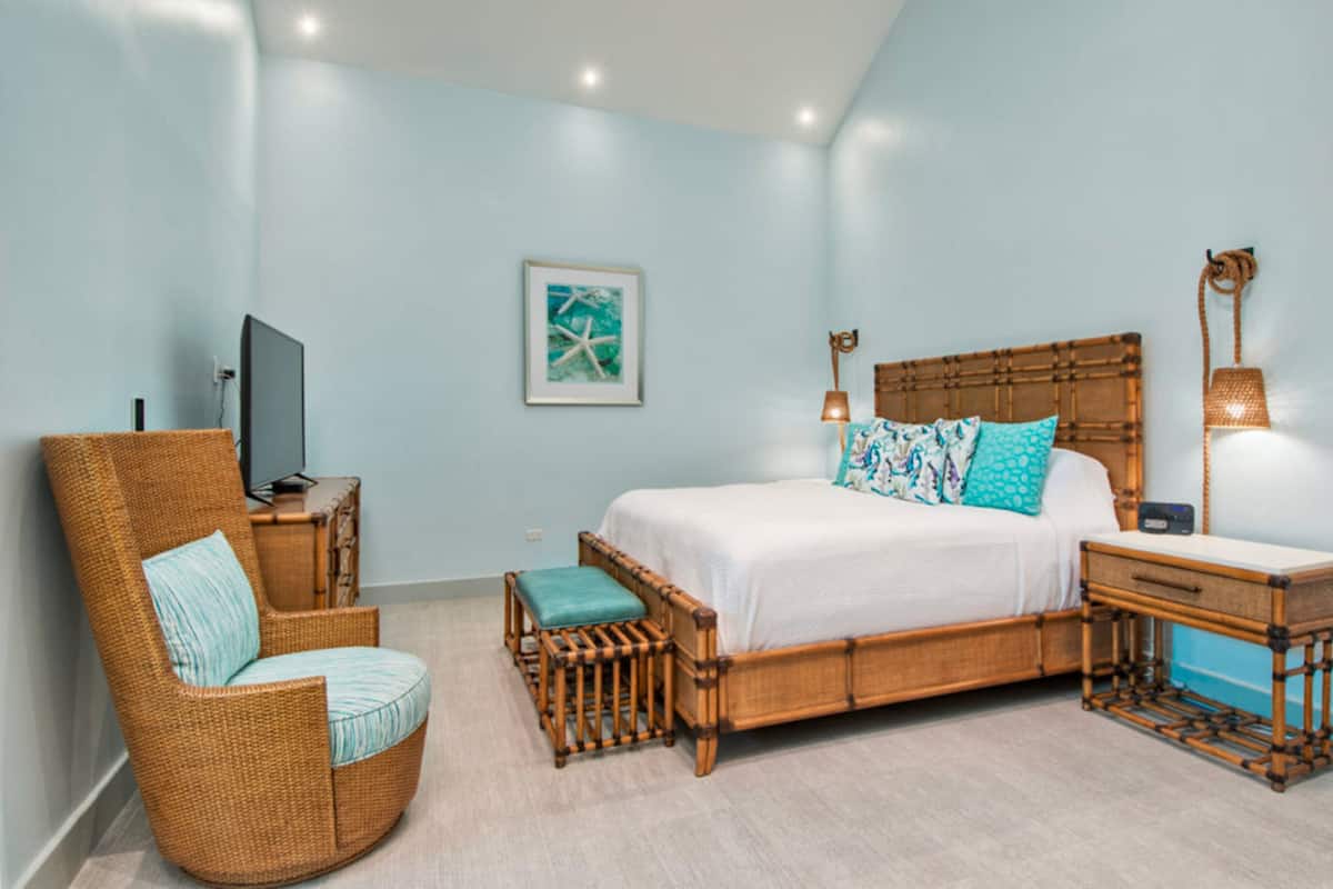 A bedroom with a bed, a wicker chair, a bedside table, a lamp, and a framed picture on the wall.