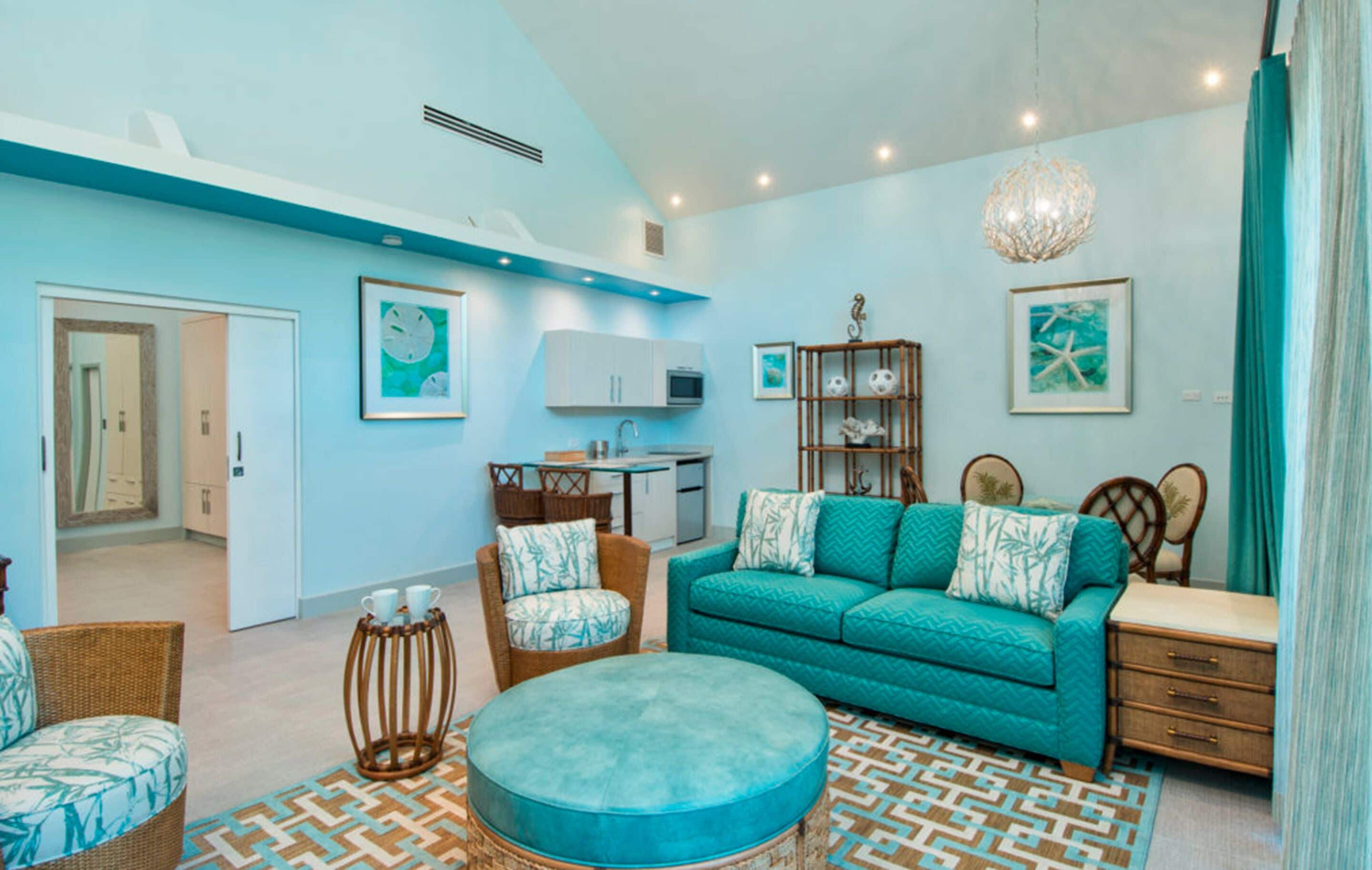 A modern living room with a teal sofa, wicker chairs, a round ottoman, and a kitchen area in the background.