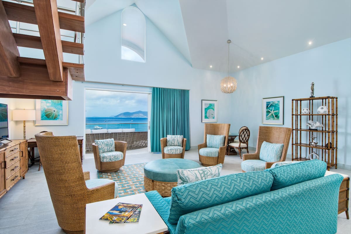 A spacious living room with wicker furniture, a large window overlooking the sea, and a ceiling with wooden beams.