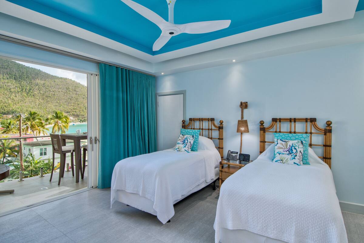 A hotel room with two beds, a ceiling fan, a balcony with a table and chairs, and a view of a mountainous landscape.
