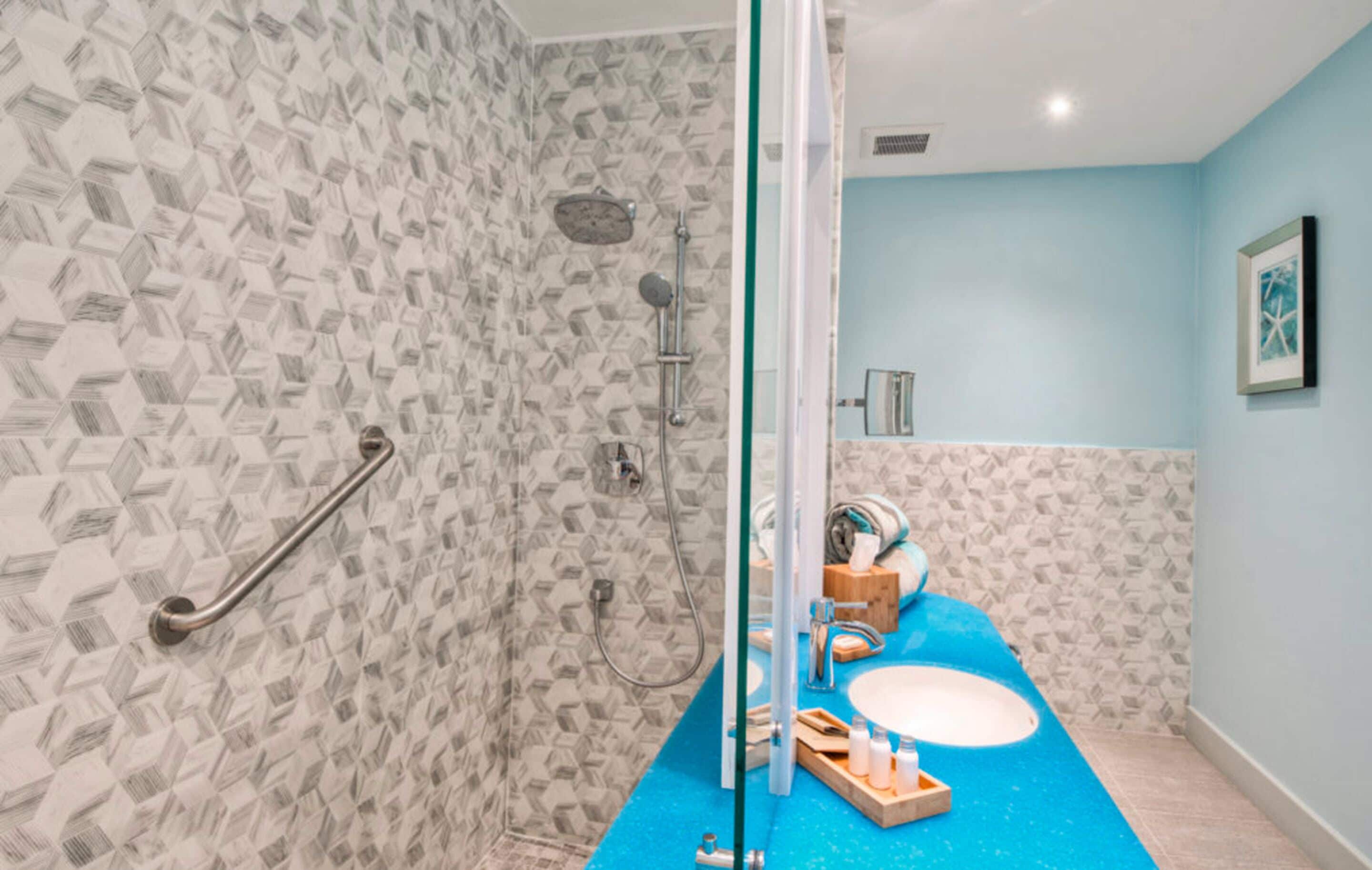 Baño con pared de patrón geométrico, ducha con cabezal de mano, lavamanos azul con espejo y toallero.