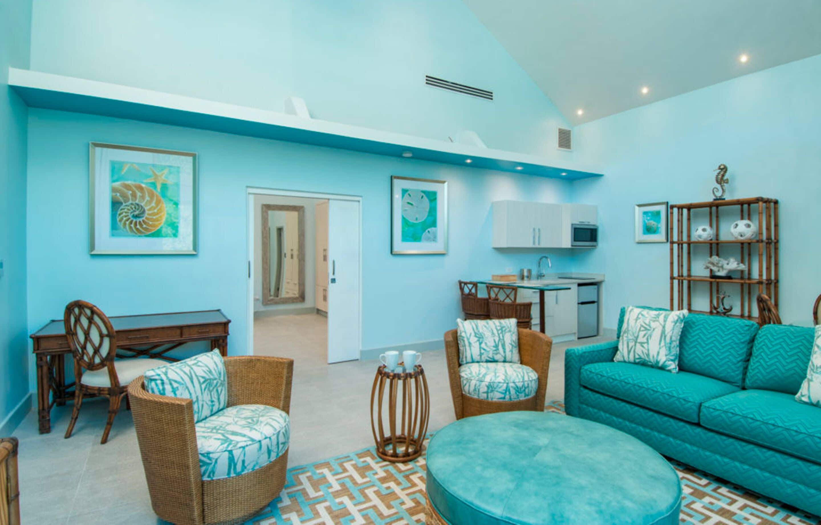 A living room with a teal sofa, wicker chairs, and a round ottoman. There is a kitchen area with cabinets and a microwave.