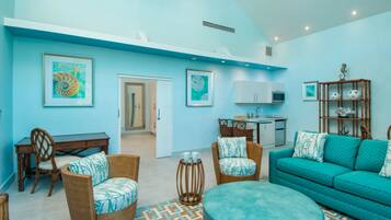 A living room with a teal sofa, wicker chairs, and a round ottoman. There is a kitchen area with cabinets and a microwave.
