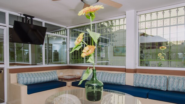 Ein Wohnzimmer mit einem Glastisch, blauen Polstersitzgelegenheiten, einer grünen Vase mit Blumen und einem Deckenventilator.