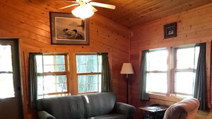 A cozy cabin interior with a wooden ceiling, a ceiling fan, a sofa, a chair, and two windows with curtains. - Welcome to Kelsey's  Cabin on the Chippewa Flowage (Hayward)