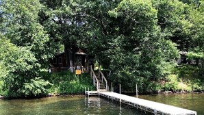 A wooden dock extending into a river, surrounded by dense foliage and a small house with a staircase leading to a deck. - Welcome to Kelsey's  Cabin on the Chippewa Flowage (Hayward)