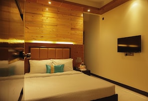 A hotel room with a wooden headboard, a flat-screen TV mounted on the wall, and a bedside table with a lamp and a phone.