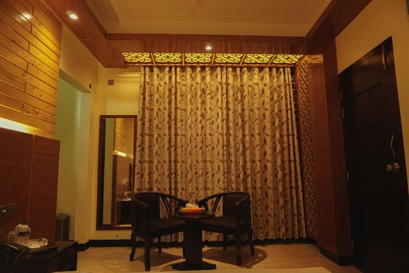 A hotel room with a wooden ceiling, a large window with patterned curtains, a round table with two chairs, and a mirror on the wall.