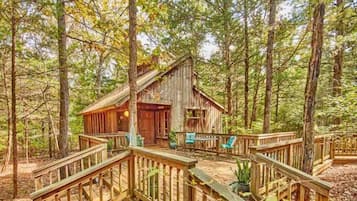 A rustic cabin with a wooden deck and a porch, surrounded by a forest.