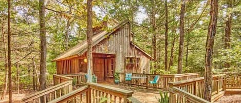 A rustic cabin with a wooden deck and a porch, surrounded by a forest.