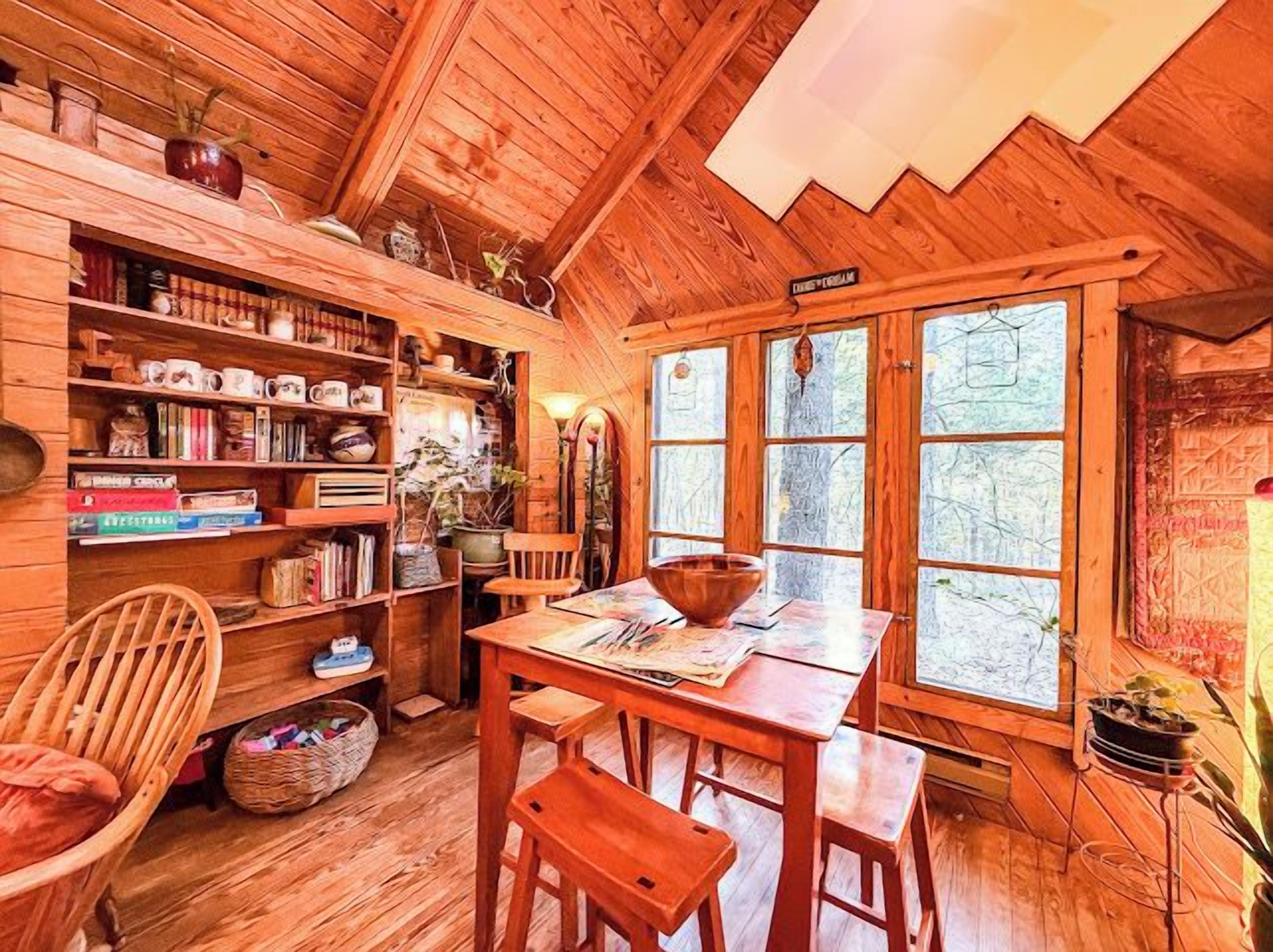 A cozy wooden cabin interior with a dining area, bookshelves, and a large window.