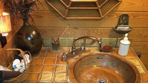 Une salle de bain avec un grand lavabo rond en cuivre, un plancher en bois, un panier en osier contenant des articles de toilette et un petit miroir.