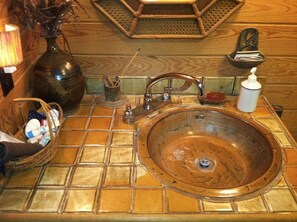 A bathroom with a large, round, copper sink, a wooden floor, a wicker basket with toiletries, and a small mirror.