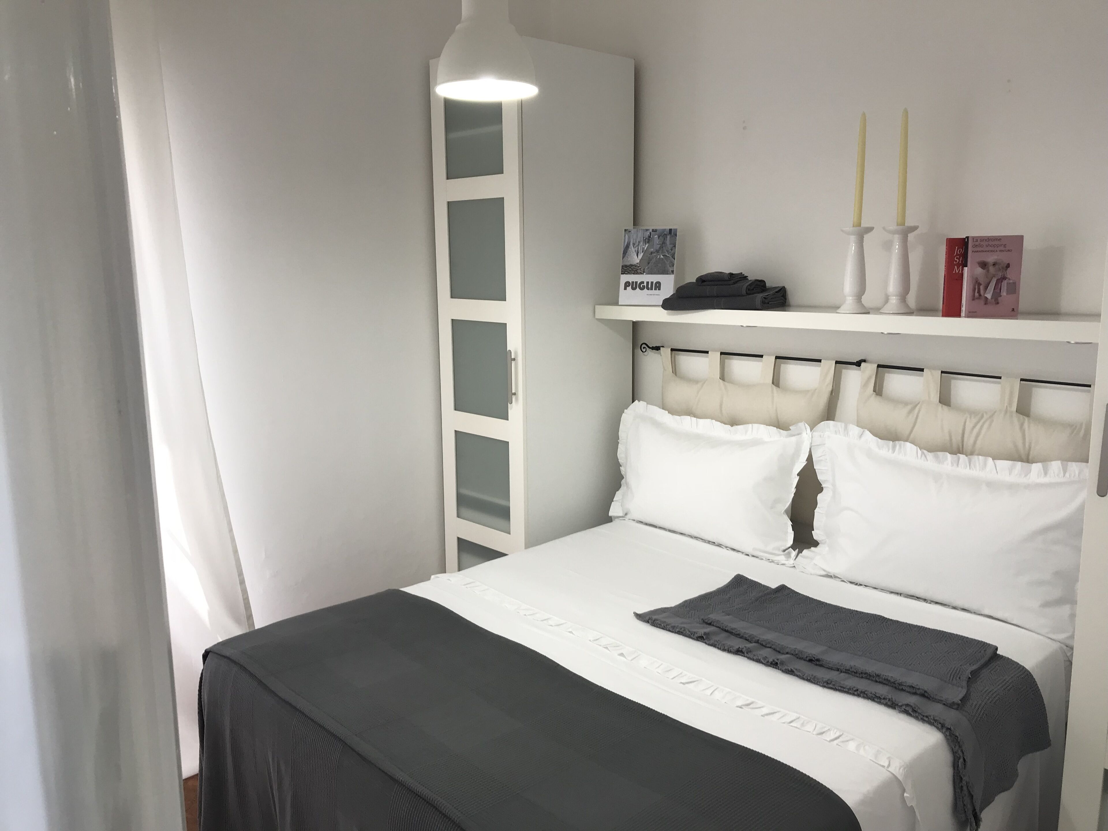 A neatly made bed with white and gray bedding, a white headboard, and a shelf above it holding a book and two candles.