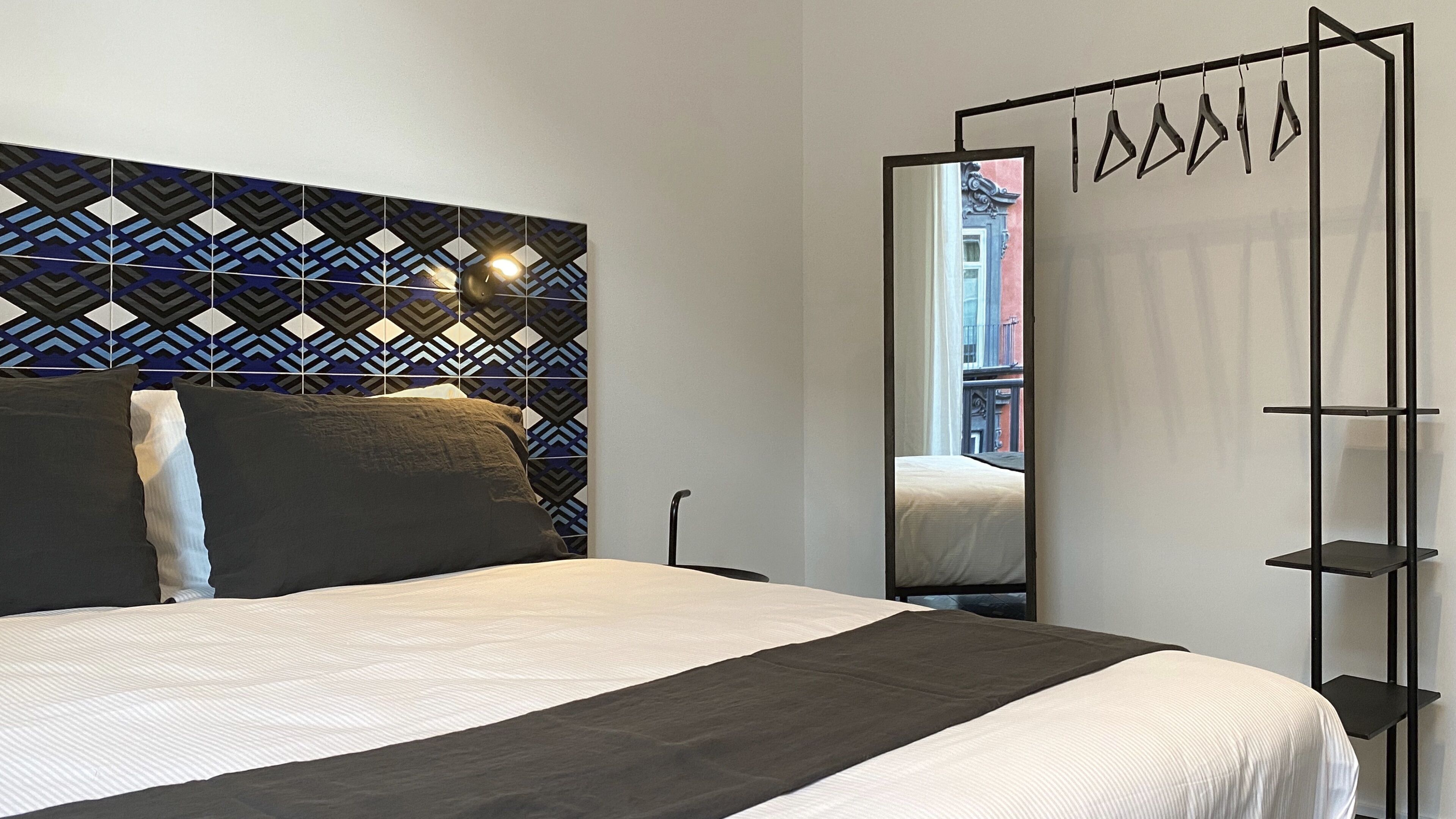 Ett modernt hotellrum med en sänggavel med geometriskt mönster, en stor spegel och en hyllsektion i svart metall.