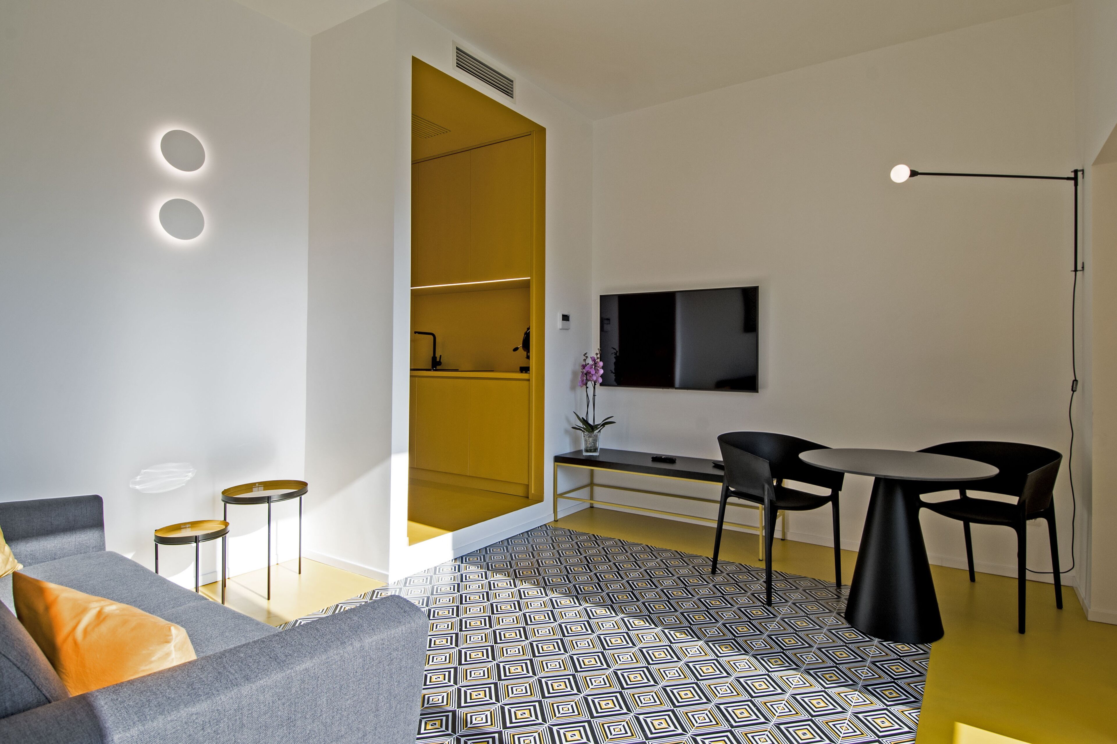 A modern living room with a sofa, coffee table, and a dining area with chairs and a table. There is a TV mounted on the wall and a floor lamp.
