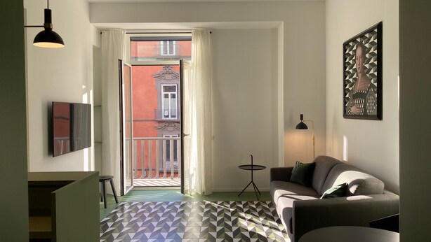 A modern living room with a sofa, a small side table, a floor lamp, a flat-screen TV, and a patterned floor. - Toraldo Suites (Naples)