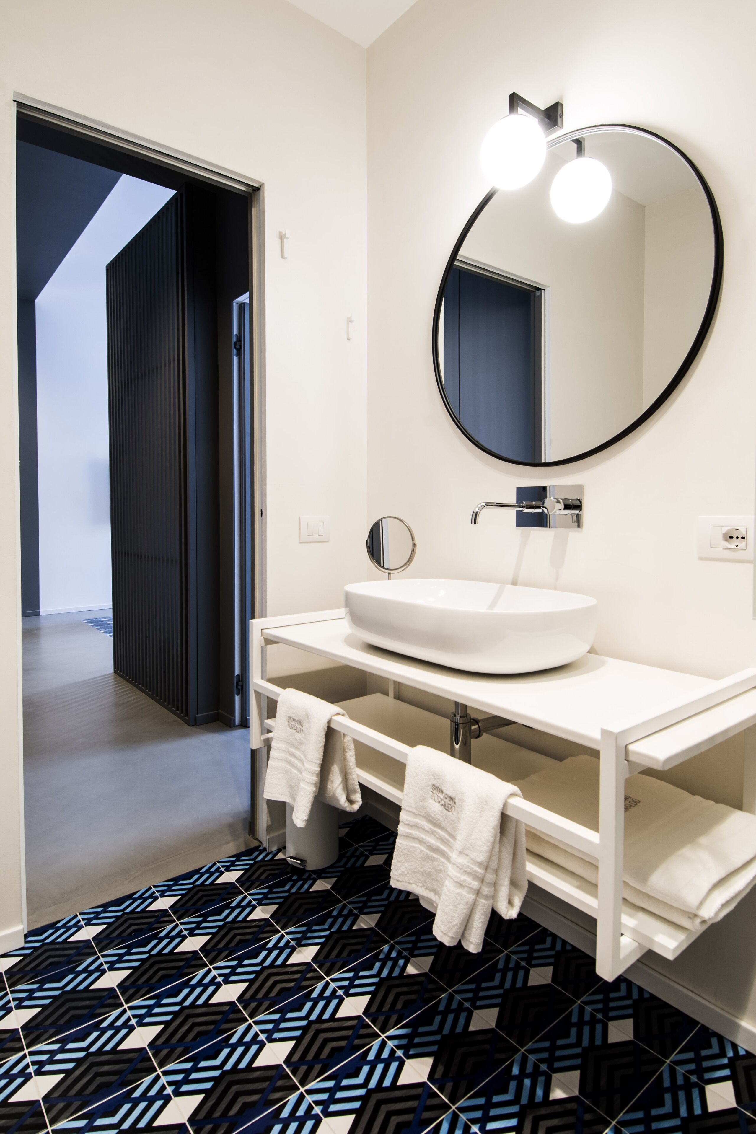 A modern bathroom with a white sink, a round mirror with lights, and patterned floor tiles.