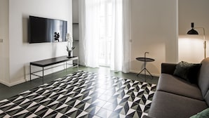 A modern living room with a black and white patterned floor, a flat-screen TV mounted on the wall, a grey sofa, and a small side table with a lamp. - Toraldo Suites (Naples)