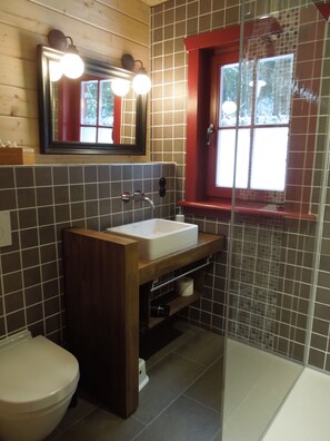 A bathroom with a wooden vanity, a white sink, a mirror with lights, a red-framed window, and a shower area. - Blockhaus Bodefall (Braunlage)