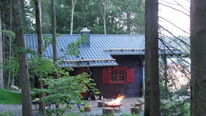A rustic cabin with a tiled roof, surrounded by trees and a fire pit in the foreground. - Blockhaus Bodefall (Braunlage)