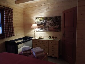 A cozy cabin room with a wooden interior, a bed with a red blanket, a dresser with drawers, a lamp, a painting of a waterfall, and a window with plaid curtains. - Blockhaus Bodefall (Braunlage)