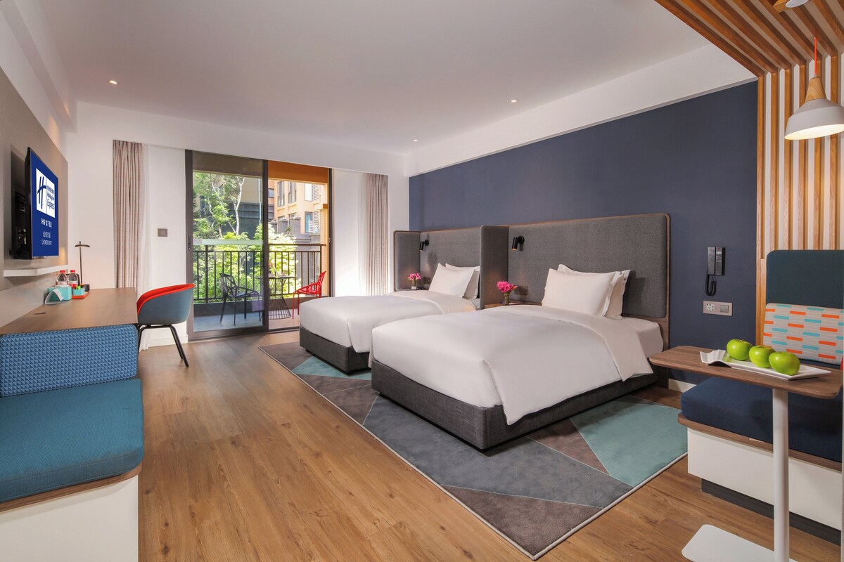 A hotel room with two beds, a desk, a chair, a TV, and a balcony with a view of buildings and trees.