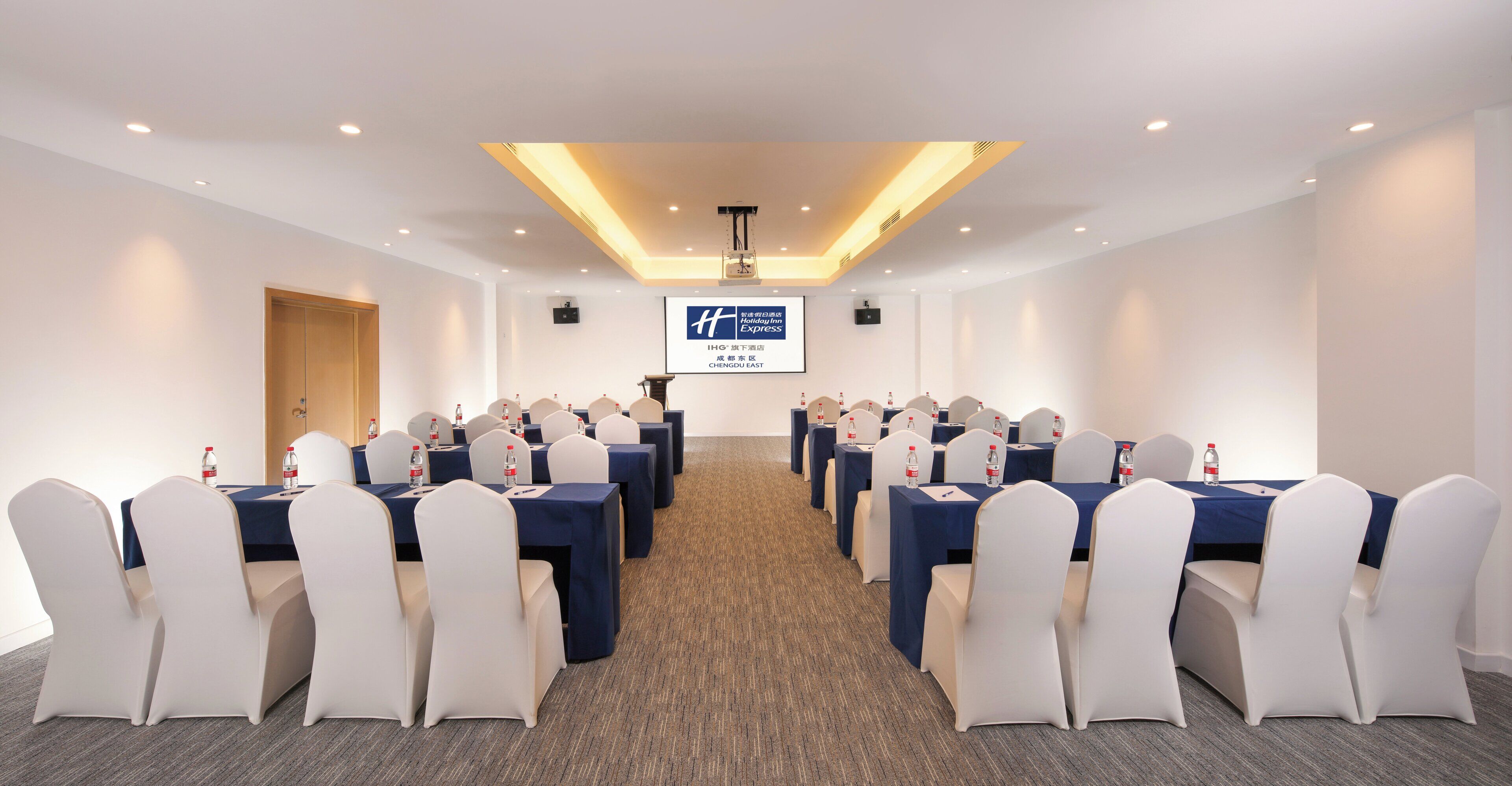 A conference room with rows of tables and chairs, a screen displaying a logo, and a door on the left.