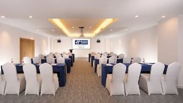 A conference room with rows of tables and chairs, a screen displaying a logo, and a door on the left.