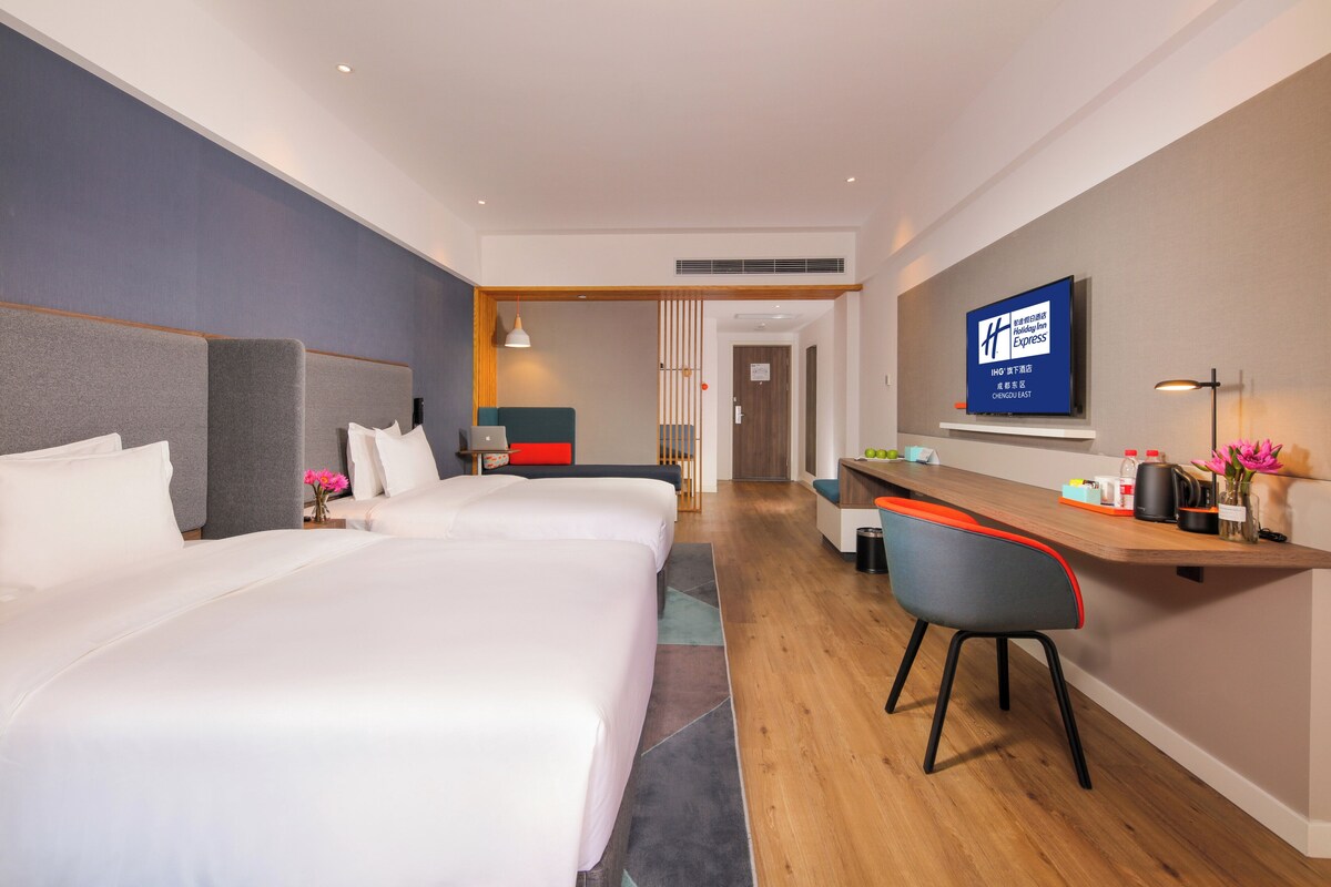 A hotel room with two beds, a desk, a chair, a TV, and a bathroom in the background.