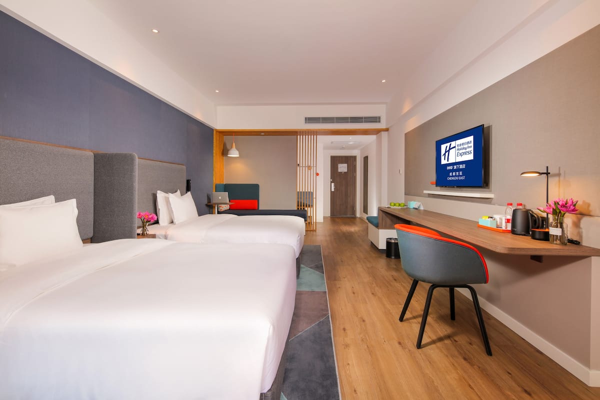 A hotel room with two beds, a desk, a chair, a TV, and a wooden floor.