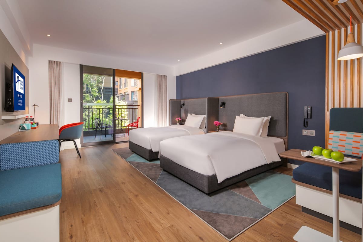 A modern hotel room with two beds, a desk, a TV, and a balcony with a view of greenery.