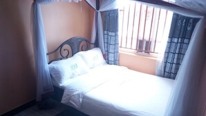 Ein Bett mit weißen Laken und einem Metall-Kopfteil, ein Fenster mit Gitter und ein Vorhang.