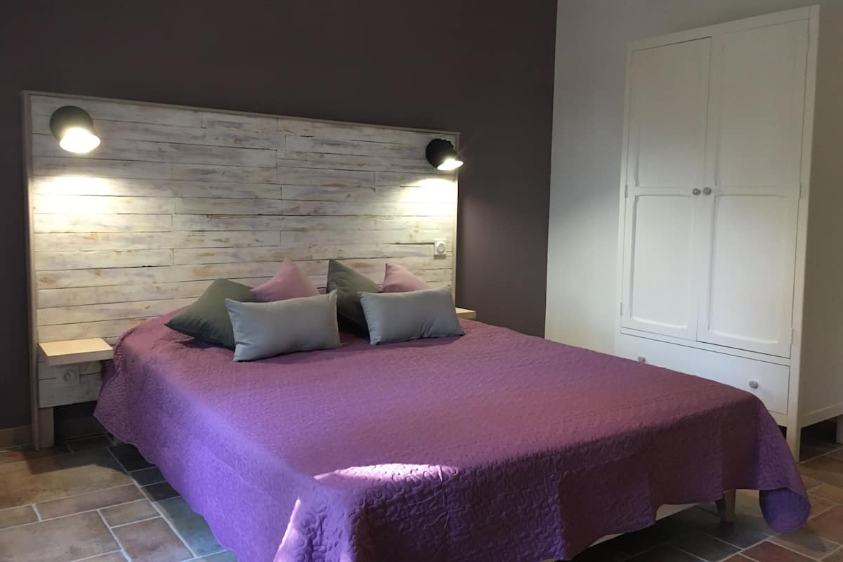 Een slaapkamer met een houten hoofdbord, een paars dekbed en twee bedlampjes.
