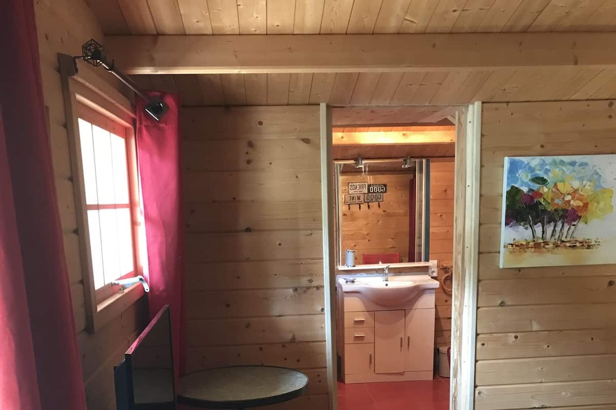 Een houten blokhut van binnen met een bed, een klein tafeltje, een wastafel en een schilderij aan de muur.