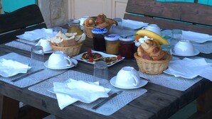 Een houten tafel gedekt met ontbijtartikelen, zoals brood, gebak en drankjes, met een uitzicht op een huis op de achtergrond.