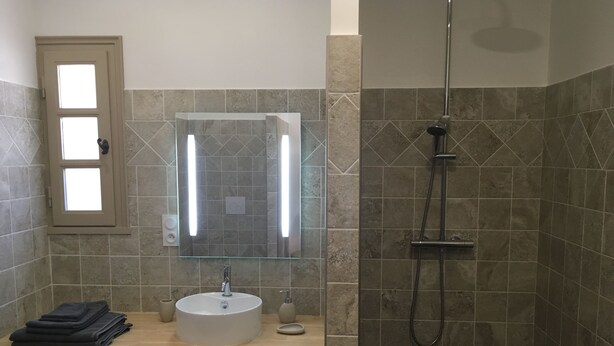 Un baño con lavamanos, espejo y ducha. Cuenta con una ventana con persianas, un perchero y una cesta.