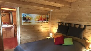 Een houten slaapkamer met een bed, kussens, een nachtkastje, een schilderij en een zichtbare badkamer door een open deur.