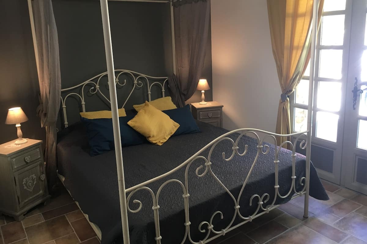 Een bed met hemelbed, voorzien van een donkerblauwe spreideken en twee kussens, met aan weerszijden nachtkastjes met lampen, in een kamer met raam en gordijnen.