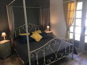 Una cama con dosel y cuatro postes, con un edredón azul oscuro y dos almohadas, flanqueada por mesitas de noche con lámparas, en una habitación con ventana y cortinas.