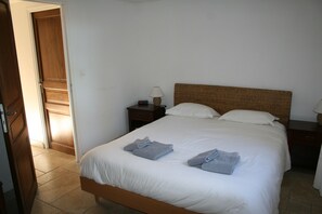 Una cama bien tendida con sábanas blancas y dos toallas dobladas, cabecera de madera, mesita de noche con lámpara y velador con reloj.